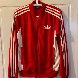 Röd Adidas jacka med vita detaljer - Säljer en snygg röd Adidas jacka med vita ränder på ärmarna och vita detaljer vid fickorna. Jackan har dragkedja och det klassiska Adidas-märket på bröstet. Perfekt för en sportig look!