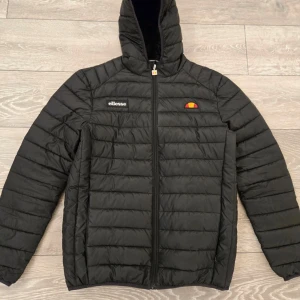 Svart pufferjacka från Ellesse - Säljer en svart pufferjacka från Ellesse med dragkedja och huva. Jackan har ett quiltat mönster och är perfekt för kyligare dagar. Den har långa ärmar och Ellesse-loggan på bröstet. Perfekt för dig som vill hålla stilen i vinter!