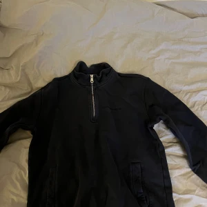 Svart tröja från J.Lindeberg - Säljer en stilren svart tröja från J.Lindeberg med halv dragkedja och broderad logga på bröstet. Perfekt för en avslappnad men snygg look. Tröjan har långa ärmar och är tillverkad i ett mjukt material.