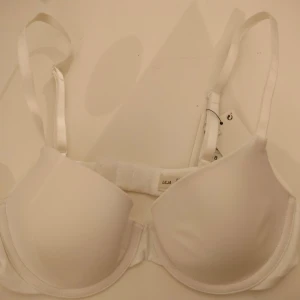 Vit T-shirt BH "Lilja" - Säljer en vit T-shirt BH "Lilja" ifrån från Lindex i storlek 70B. Den har släta kupor och bygel för extra stöd. Tillverkad i en återvunnen polyamidblandning för en mjuk och bekväm passform. Perfekt för dagligt bruk. Helt oanvänd, prislapp sitter kvar.