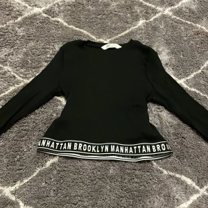 Svart långärmad topp från H&M - Snygg svart långärmad topp från H&M med texten 'Brooklyn Manhattan' längs nederkanten. Perfekt för en cool och avslappnad stil. Tillverkad i ett ribbat material som ger en skön passform.