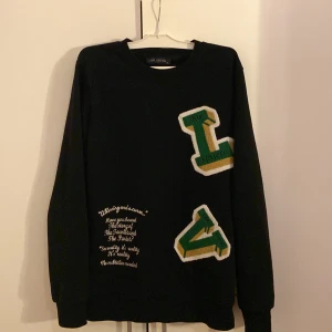 Svart långärmad tröja från Louis Vuitton - Snygg svart långärmad tröja från Louis Vuitton med broderade bokstäver i grönt och guld. Textdetaljer på framsidan ger en unik touch. Perfekt för dig som vill ha något stilrent men ändå iögonfallande.