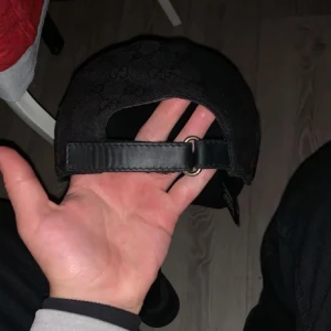 Svart gg keps med mönster  - Snygg svart keps från Gucci med diskret mönster och justerbart bälte baktill. Kepsen har en klassisk design med röd och grön randdetalj på sidan. Perfekt för att ge din outfit en lyxig touch.