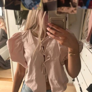 Ljusrosa blus med knytning - Säljer en söt ljusrosa blus med puffiga kortärmar och knytning framtill. Helt nyskick kostar orginellt 500kr