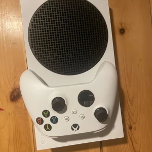 Vit Xbox Series S med handkontroll - Säljer en vit Xbox Series S tillsammans med en matchande vit handkontroll. Konsolen har en kompakt design med en stor svart ventilationsgrill på toppen. Perfekt för gaming med hög prestanda i ett litet format. Inkluderar flera anslutningsportar på baksidan.