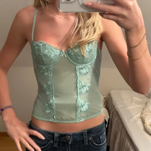 Mintgrön korsettopp - Snygg mintgrön korsettopp med spetsdetaljer och justerbara axelband. Toppen har en figurnära passform och är perfekt för att ge en stilren look. Den har en vacker spetsdesign framtill och en slät baksida.