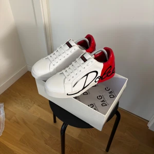 Dolce & Gabbana Portofino - Snygga vita sneakers från Dolce & Gabbana med röd häl och svart textdetalj. Skorna har snörning och en stilren design med märkets logga på sidan och hälen. Perfekta för att ge en touch av lyx till din outfit.