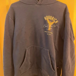 Mörkblå hoodie från Hoodrich - Säljer en mörkblå hoodie från Hoodrich med ett stort tryck av en ängel och texten 'From Nothing To Something' i guld på både fram- och baksidan. Hoodien har en stor ficka framtill och är perfekt för en avslappnad stil.