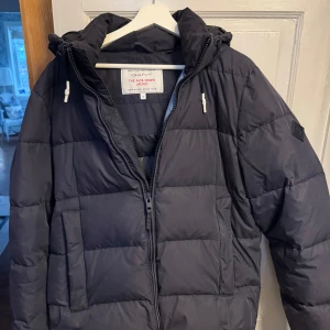 Mörkblå dunjacka från GANT - Säljer en mörkblp dunjacka från GANT, modell 'The Alta Down Jacket'. Jackan har en dragkedja framtill och en huva med dragsko. Perfekt för kyliga dagar med sin quiltade design och praktiska fickor. Nypris 2600kr.