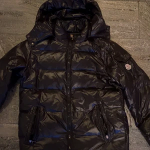 Moncler maya  - Säljer en svart pufferjacka från Moncler med huva och dragkedja. Jackan har ett quiltat mönster och en broderad logotyp på ärmen. Perfekt för kyliga dagar.