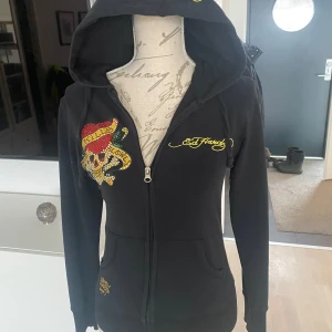 Svart hoodie från Ed Hardy - Snygg svart hoodie från Ed Hardy med färgglatt dödskallemotiv och texten 'Love Kills Slowly' i strass på framsidan och baksidan. Hoodien har dragkedja och långa ärmar, perfekt för en cool och avslappnad stil. Haft denna tröja i år och det är en stor favorit (tar inga scam bud) står L men är en S/XS