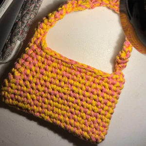 Gul och rosa virkad handväska - Säljer en färgglad handväska i virkat material med ett unikt mönster i gult och rosa. Väskan har ett flätat handtag som ger en extra touch av stil. Perfekt för att liva upp vilken outfit som helst!
