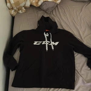 Svart hoodie från CCM - Säljer en svart hoodie från CCM med vit logga på bröstet. Den har en klassisk design med känguruficka och justerbar huva med snören. Perfekt för en avslappnad stil.