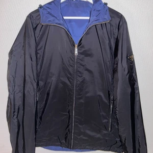 Svart vindjacka från Prada - Snygg svart vindjacka från Prada med blått foder och dragkedja framtill. Jackan har en klassisk design med långärmade ärmar och en diskret logga på ärmen. Perfekt för en stilren look.