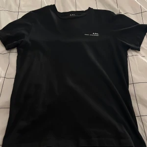 Svart t-shirt från A.P.C. - Snygg svart t-shirt från A.P.C. med tryck på bröstet. Perfekt för en stilren look. Tillverkad i 100% bomull. T-shirten är använd men i bra skick.  Storlek S. 
