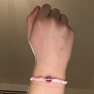 Rosa armband med smiley - Charmigt rosa armband med en glad smiley-berlock i mitten. Armbandet är flätat och justerbart, perfekt för att ge en lekfull touch till din stil.