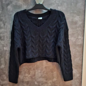 Svart stickad tröja med v-ringning - Snygg svart stickad tröja med v-ringning och kabelstickat mönster. Tröjan är croppad och har långa ärmar, perfekt för en stilren look. Aldrig använd