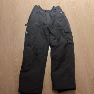 Svarta täckbyxor med dragkedja - Snygga svarta täckbyxor med praktiska dragkedjor och en cool logga på benet. Perfekta för kalla dagar och utomhusaktiviteter. Byxorna har en bekväm passform och elastisk midja för extra komfort.