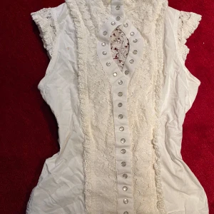 vit blus med knäppning - säljer min favoritblus för den passar inte mig längre! den har unik knäppning hela vägen från halsen ned, med undantag från en bit ovan bysten. genomskinlig i ryggen med underbar rosettdetalj. bomull, timjan design. stl XS