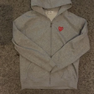 Grå hoodie från Comme des Garçons Play - Säljer en snygg grå hoodie från Comme des Garçons Play i storlek M. Den har en dragkedja framtill och ett ikoniskt rött hjärta med ögon på bröstet. Perfekt för en casual look eller mysiga dagar hemma. 🖤