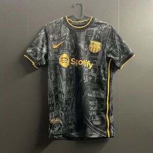 Svart och guld FC Barcelona fotbollströja - Säljer en snygg svart FC Barcelona tröja med guldiga detaljer och tryck. Den har korta ärmar och ett coolt mönster med stadsmotiv. Perfekt för alla Barca-fans! Tröjan är i mycket bra skick och har både Nike och Spotify loggor. Passa på att köpa denna unika tröja!