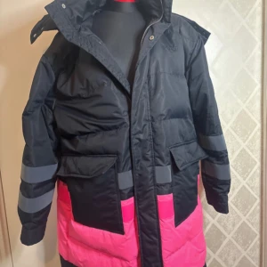 Svart och rosa dunjacka från Svea - Säljer en snygg och varm dunjacka från Svea i svart och rosa med grå detaljer. Jackan har långa ärmar och praktiska fickor framtill. Perfekt för kalla dagar med sin pufferdesign och huva.