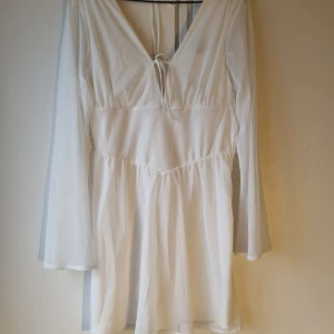 Vit klänning med korsettliv - Elegant vit klänning med v-ringning och knytband framtill, samt öppen rygg. Långärmad design med lätt och flödande material som ger en luftig känsla. Perfekt för varmare dagar.