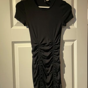 Svart klänning från H&M Divided - Snygg svart klänning från H&M Divided i storlek XS. Klänningen har korta ärmar och en åtsittande passform med rynkade detaljer längs sidorna 