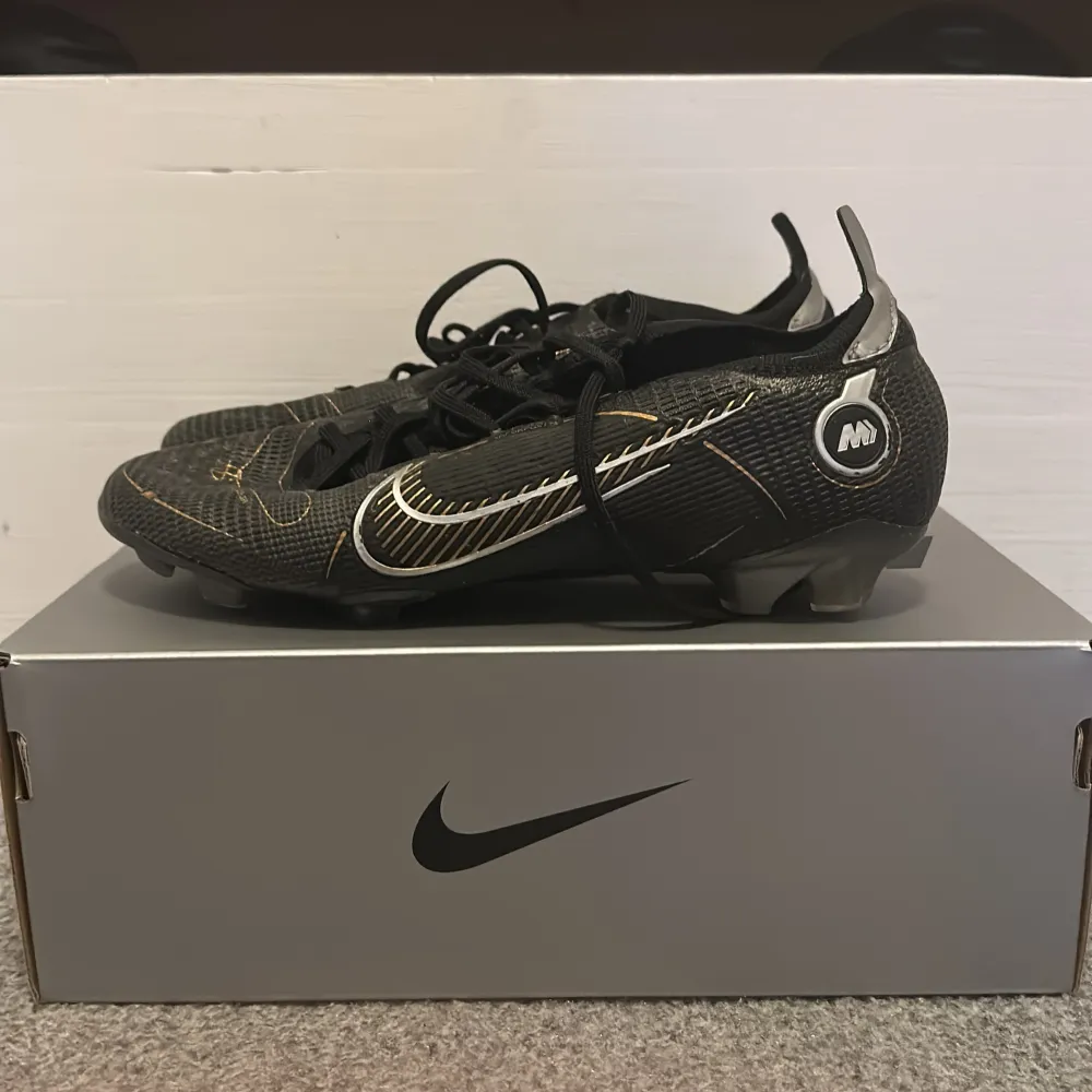 Snygga svarta fotbollsskor från Nike med vita och guldiga detaljer. Skorna har snörning och är designade för optimal hastighet och kontroll på planen. Perfekta för fotbollsspelare som vill ha stil och funktionalitet. Mitt pris 299 nytt pris 3299. Skor.