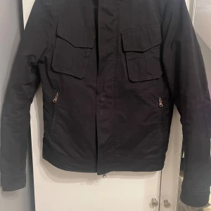 Svart jacka från Oscar Jacobson - Snygg svart jacka från Oscar Jacobson med dragkedja och knappar. Jackan har två bröstfickor med lock och två sidofickor med dragkedja. Perfekt för en stilren look. Storlek S.