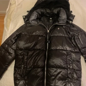 Svart pufferjacka från G-Star RAW - Säljer en snygg svart pufferjacka från G-Star RAW. Jackan har en dragkedja framtill, en hög krage och luva som går att ta av. Perfekt för kalla vinterdagar. Den är i mycket bra skick och passar både till vardags och lite finare tillfällen.