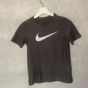 Grå Dri-FIT t-shirt från Nike - Säljer en grå Dri-FIT t-shirt från Nike i mycket bra skick. Perfekt för träning med sin fukttransporterande teknik. Den har en klassisk Nike-logga i vitt på bröstet och är kortärmad.