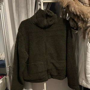 Olivgrön teddyjacka från H&M - Mysig olivgrön teddyjacka från H&M med dragkedja framtill. Jackan har en hög krage och två praktiska fickor. Perfekt för kyliga dagar när du vill hålla dig varm och bekväm. Finns i M också!