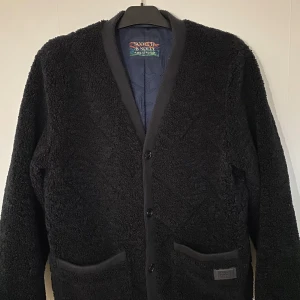 Svart teddyjacka från Scotch & Soda - Mysig svart teddyjacka från Scotch & Soda, perfekt för kyliga dagar. Jackan har en v-ringad design med knappar framtill och två praktiska fickor. Den är i mycket bra skick och ger en stilren look. Passar både till vardags och lite finare tillfällen.