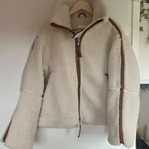 Beige teddyjacka med dragkedja - Mysig beige teddyjacka med dragkedja och bruna detaljer. Perfekt för kyliga dagar med sin fluffiga och varma design. Slutsåld på H&M.