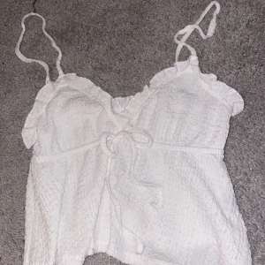 Vit ärmlös topp med volangdetaljer - Säljer en söt vit topp med smala axelband och volangdetaljer. Toppen har en knytning framtill. Materialet är lätt och luftigt, vilket gör den bekväm att bära. Det finns inga skador eller fläckar🤍säljer pga att det inte används längre🫶🏻 