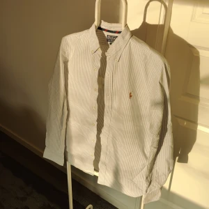 Randig skjorta från Polo Ralph Lauren - Säljer en stilren och klassisk randig skjorta från Polo Ralph Lauren i slim fit. Skjortan är vit med blå ränder och har en broderad logga på bröstet. Perfekt för både vardag och fest! Den är i mycket bra skick och har långa ärmar med knappar.