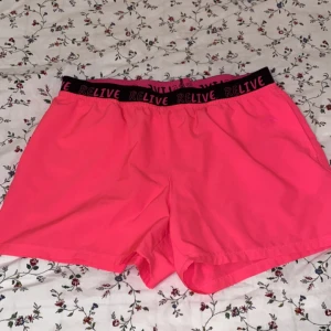 Rosa shorts från Crivit - Säljer ett par knallrosa shorts från Crivit i storlek M. De har en elastisk midja med texten 'RELIVE' i svart. Perfekta för sommarens alla aktiviteter, oavsett om det är träning eller bara hänga med vänner. Superbekväma och i bra skick!