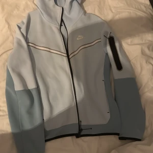 Ljusblå hoodie från Nike - Säljer en ljusblå hoodie från Nike med dragkedja och huva. Jackan har en stilren design med svarta detaljer och en ficka på ärmen. Perfekt för en sportig look. Äkta och väldigt sällsynt.