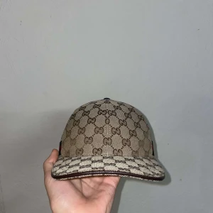 Beige keps från Gucci - Snygg beige keps från Gucci med det ikoniska GG-mönstret. Kepsen har en justerbar rem baktill och en grön och röd randdetalj på sidan. Perfekt för att ge din outfit en lyxig touch. Kepsen är endast använd 3 gånger och är i nyskick. Pris kan diskuteras vid snabbaffär 