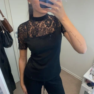 Svart spetsblus - Elegant svart blus med spetsdetaljer på överdelen och korta ärmar. Perfekt för en stilren look med en touch av romantik. Blusen har en hög hals och en liten knappöppning i nacken. Blusen är M men känns som S.