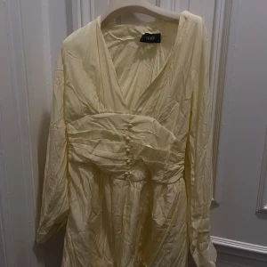 Ljusgul klänning från Cider - Elegant ljusgul klänning från Cider med långa ärmar och v-ringning. Klänningen har knappar framtill och ett fint rynkat parti vid midjan som ger en smickrande passform. Perfekt för vårens och sommarens tillfällen.