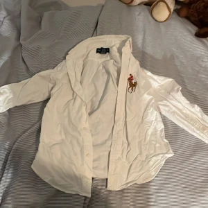Vit skjorta från Ralph Lauren - Säljer en klassisk vit skjorta från Ralph Lauren med en snygg broderad polospelare på bröstet. Skjortan har långa ärmar och knappar framtill. Perfekt för både vardag och fest! 🐎