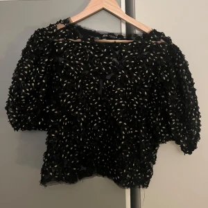 Svart blus med mönster från Zara - Säljer en unik svart blus från Zara med ett coolt mönster i guld. Blusen har puffiga ärmar och en luftig passform, perfekt för en festlig kväll. I storlek L men liten i storlek så passar också M