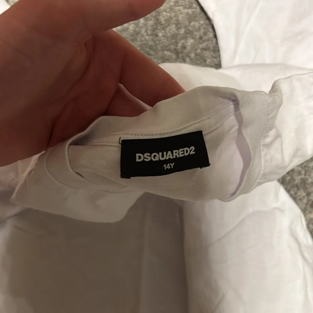 Säljer en snygg vit t-shirt från Dsquared2 i storlek 14Y. Den har en stilren design med ett svart lönnlöv och text på bröstet. Perfekt för en casual look eller att styla upp med en jacka. T-shirten är i bra skick och gjord av mjuk bomull. Passar perfekt till sommaren! 🌟köpt på kids brand store. T-shirts.