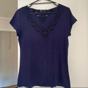 Mörkblå topp från Lindex - Snygg mörkblå topp från Lindex med v-ringad spetsdetalj. Perfekt för både vardag och fest. Den har korta ärmar och är gjord i ett mjukt material som känns skönt mot huden.