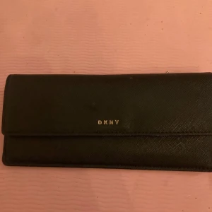 Svart plånbok från DKNY - Elegant svart plånbok från DKNY med stilren design. Plånboken har en praktisk dragkedja på baksidan och flera fack inuti för kort och kontanter. Knappt använd Nypris 800 kr  ev frakt betalas av köparen