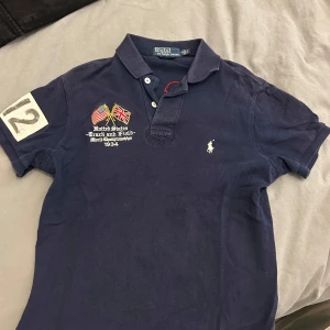 Mörkblå piké från Ralph Lauren - Snygg mörkblå pikétröja från Ralph Lauren med broderade flaggor och text på framsidan. Tröjan har korta ärmar och en klassisk krage med knappar. På baksidan finns stora tryck med 'U.S.A.' och siffran '012'. Perfekt för en sportig look!