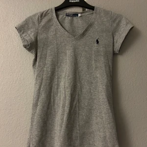 Grå t-shirt från Polo - Säljer en klassisk grå t-shirt från Polo i 100% bomull. Den har en v-ringad hals och korta ärmar, perfekt för vardagsbruk. Den lilla loggan på bröstet ger en stilren touch. Passar bra till jeans eller shorts för en avslappnad look.