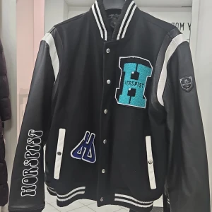 Svart och vit bomberjacka från Horspist - Säljer en snygg svart och vit bomberjacka från Horspist i storlek XL. Jackan har coola broderade detaljer och tryck, med dragkedja och knappar framtill. Perfekt för höst och vår med sin quiltade insida. Den har långa ärmar och en unik design på ryggen. Passar perfekt för dig som vill sticka ut! Jackan är lite mindre I storleken, motsvarar en L storlek.